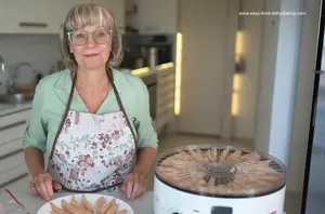 Smiling woman in floral apron standing beside a round white food dehydrator with the top tray removed, showing dehydrated chicken slices now cooling on a nearby plate.