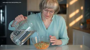 woman pouring filtered water from a measuring jug into a bowl of dried vegetables on a kitchen counter.