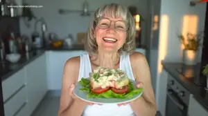 Smiling woman in glasses holding a plate with a chicken salad sandwich on lettuce and tomato in a bright kitchen