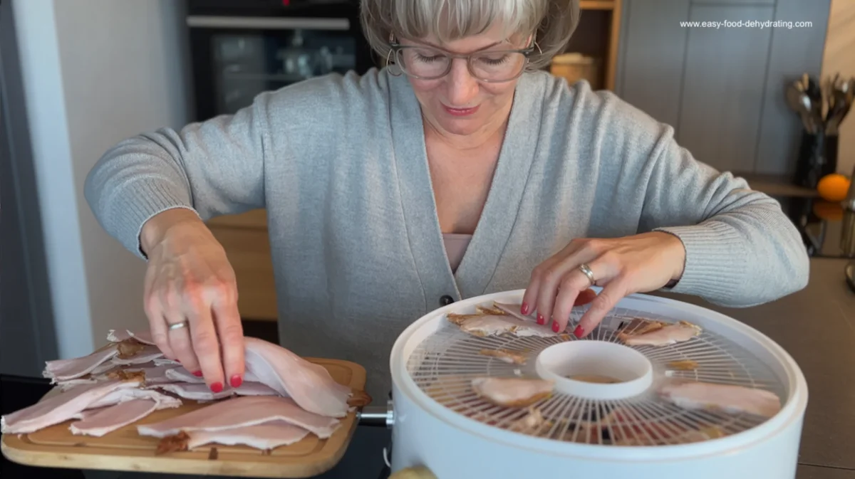 Woman slicing cooked turkey breast and arranging strips on a round white food dehydrator tray, preparing homemade dried turkey for safe long-term storage.