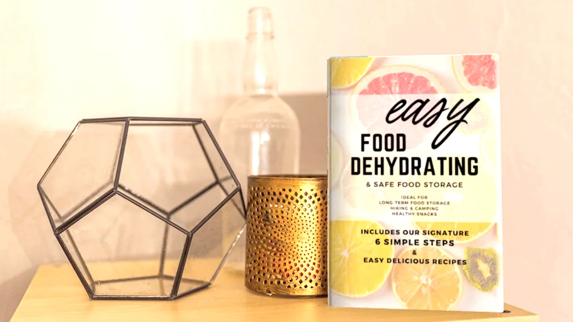 Easy Food Dehydrating book displayed on a wooden shelf beside modern home decor, including a geometric glass frame, gold candle holder, and clear bottle.