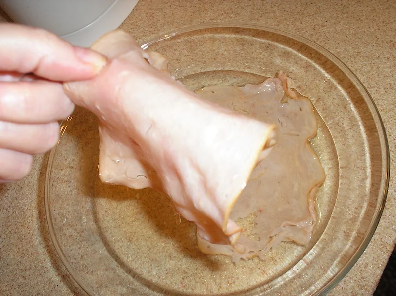 Hand lifting a slice of dehydrated deli turkey from a bowl of cold water during rehydration.