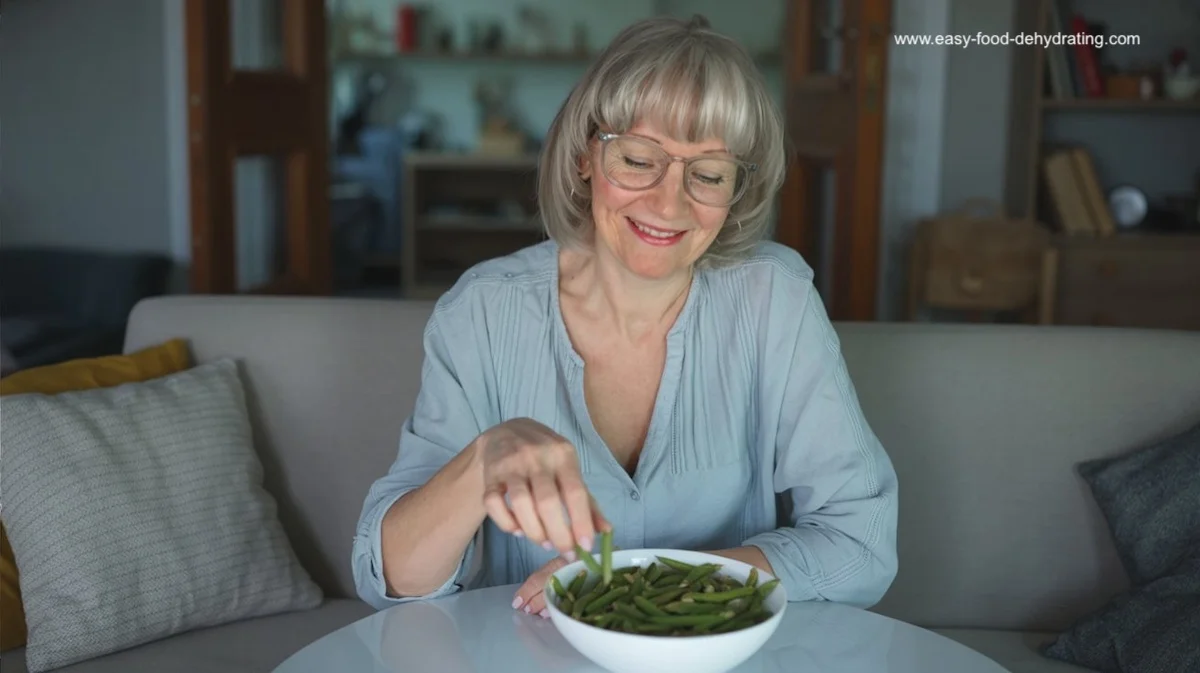Susan sits in her living room, picking up a crispy dehydrated green bean snack from a white bowl on the table in front of her.